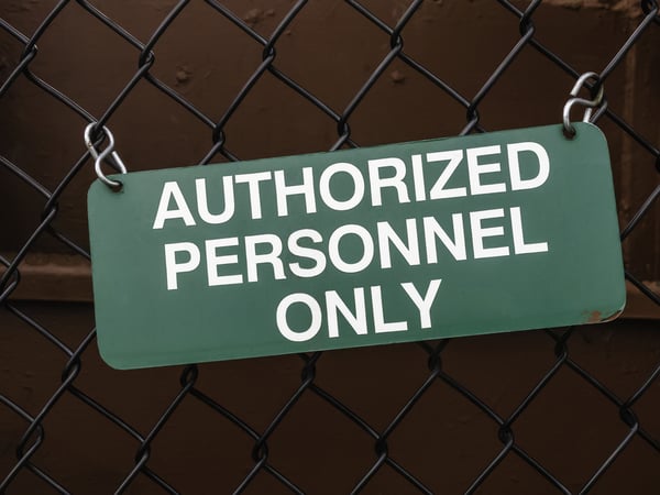 Light green outdoor sign, slightly crooked, on black chain-link fence by dark red steel wall AUTHORIZED PERSONNEL ONLY